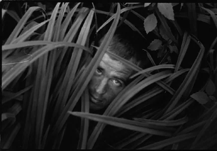 Black and white photograph: a face half hidden, looking out through blades of tall grass